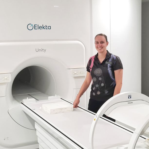 ֱ Alumni Ari Brodmann smiling and standing next to the MR-Linac machine that provides targeted radiation treatment. 